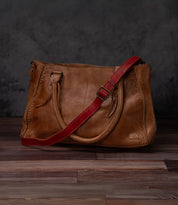A timeless Bed|Stü Crossbody Strap in brown leather with decorative stitching, two handles, and a detachable red adjustable leather strap for crossbody wear—styled on a wooden floor against a dark textured background.