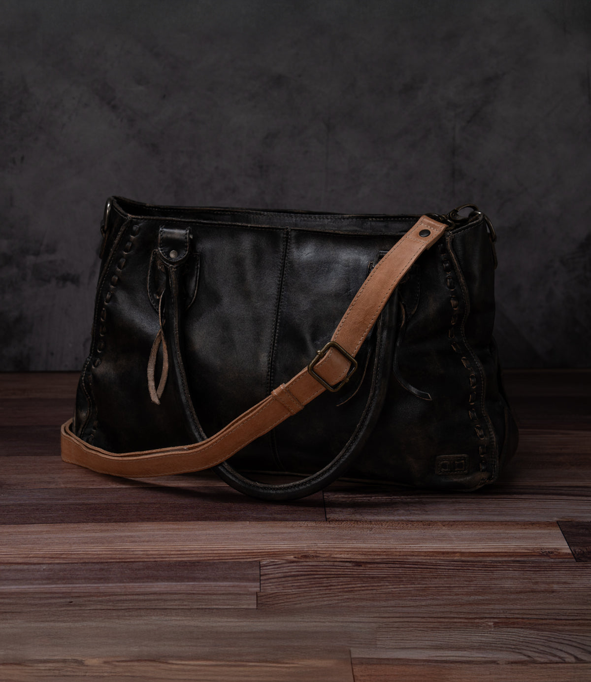 A Bed|Stü Crossbody Strap in black leather, featuring an adjustable leather strap for versatile crossbody styling, rests on a wooden floor against a dark gray textured background, embodying the classic spirit of Bed|Stü bags.
