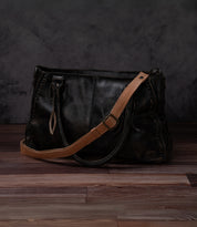 A Bed|Stü Crossbody Strap in black leather, featuring an adjustable leather strap for versatile crossbody styling, rests on a wooden floor against a dark gray textured background, embodying the classic spirit of Bed|Stü bags.