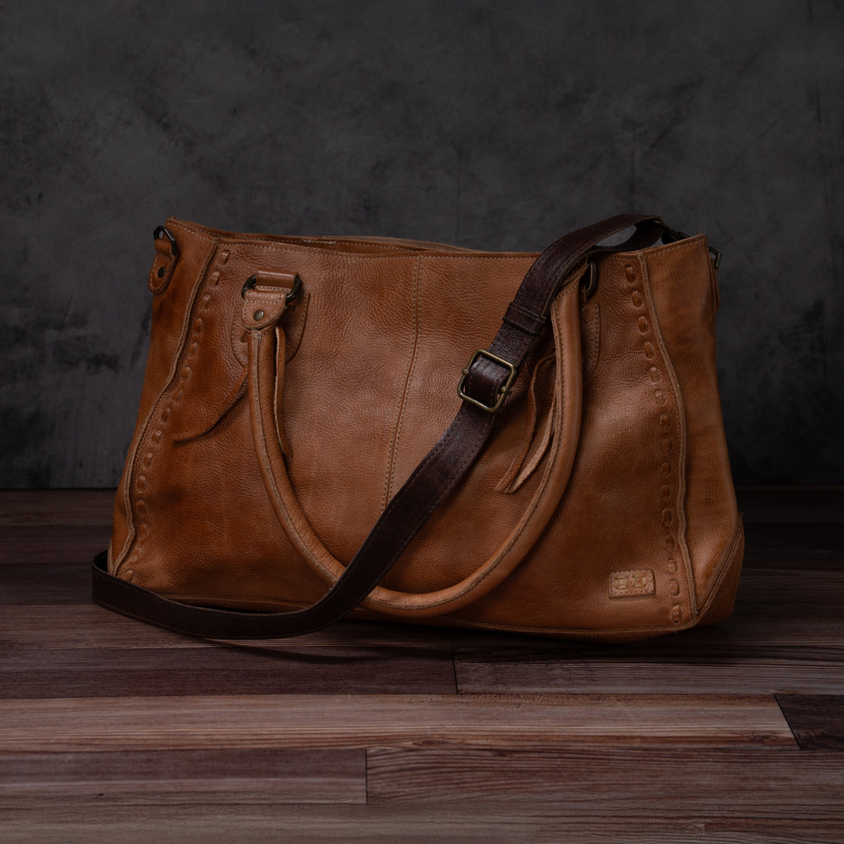 A classic Bed|Stü brown leather handbag featuring dual handles and a dark brown adjustable Crossbody Strap is showcased on a wooden floor against a dark, textured backdrop, embodying the brand's timeless craftsmanship.