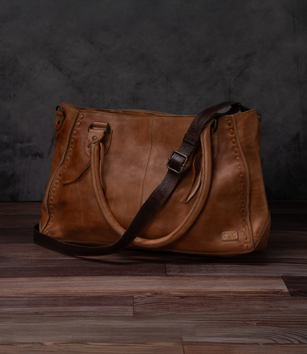 A classic Bed|Stü brown leather handbag featuring dual handles and a dark brown adjustable Crossbody Strap is showcased on a wooden floor against a dark, textured backdrop, embodying the brand's timeless craftsmanship.