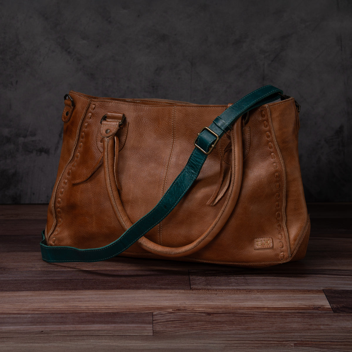 Bed|Stü Crossbody Strap in brown leather with decorative stitching, featuring dual handles and an adjustable strap for versatile crossbody wear, displayed on a dark wooden surface against a dark textured background.