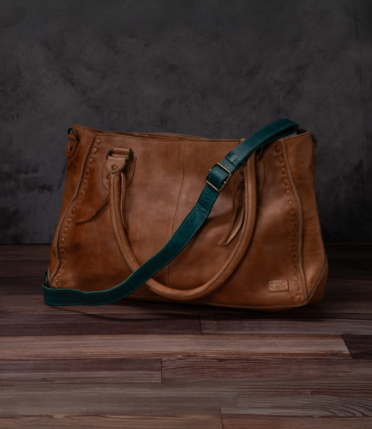 Bed|Stü Crossbody Strap in brown leather with decorative stitching, featuring dual handles and an adjustable strap for versatile crossbody wear, displayed on a dark wooden surface against a dark textured background.