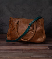 Bed|Stü Crossbody Strap in brown leather with decorative stitching, featuring dual handles and an adjustable strap for versatile crossbody wear, displayed on a dark wooden surface against a dark textured background.