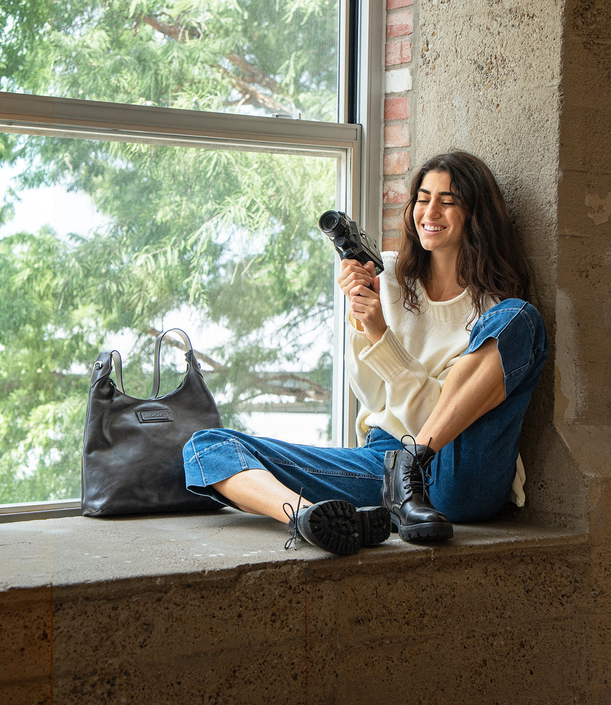 A person wearing a white sweater, jeans, and black lace-up boots sits in a window sill holding a vintage video camera, with a black leather Bed|Stü Dallas LTC shoulder bag beside them.
