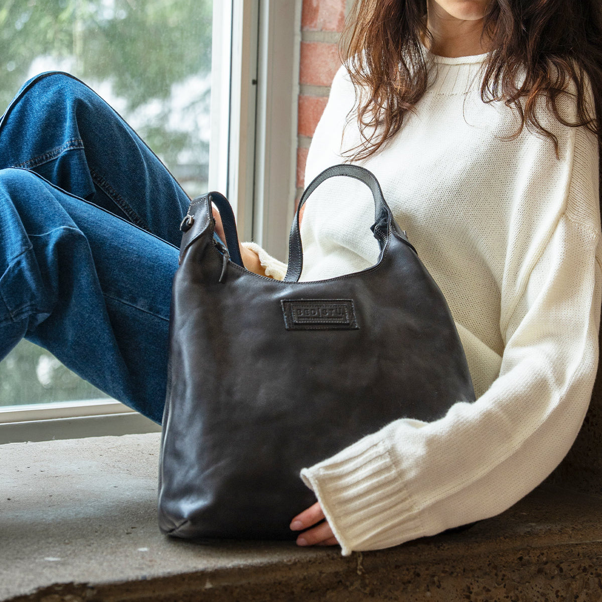 A woman in a white sweater sits by a window, holding a dark leather Bed|Stü Dallas LTC shoulder bag.