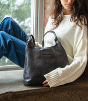 A woman in a white sweater sits by a window, holding a dark leather Bed|Stü Dallas LTC shoulder bag.