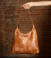 A hand holds a tan leather Bed|Stü Dallas LTC hobo bag with a zipper closure and branded leather patch.
