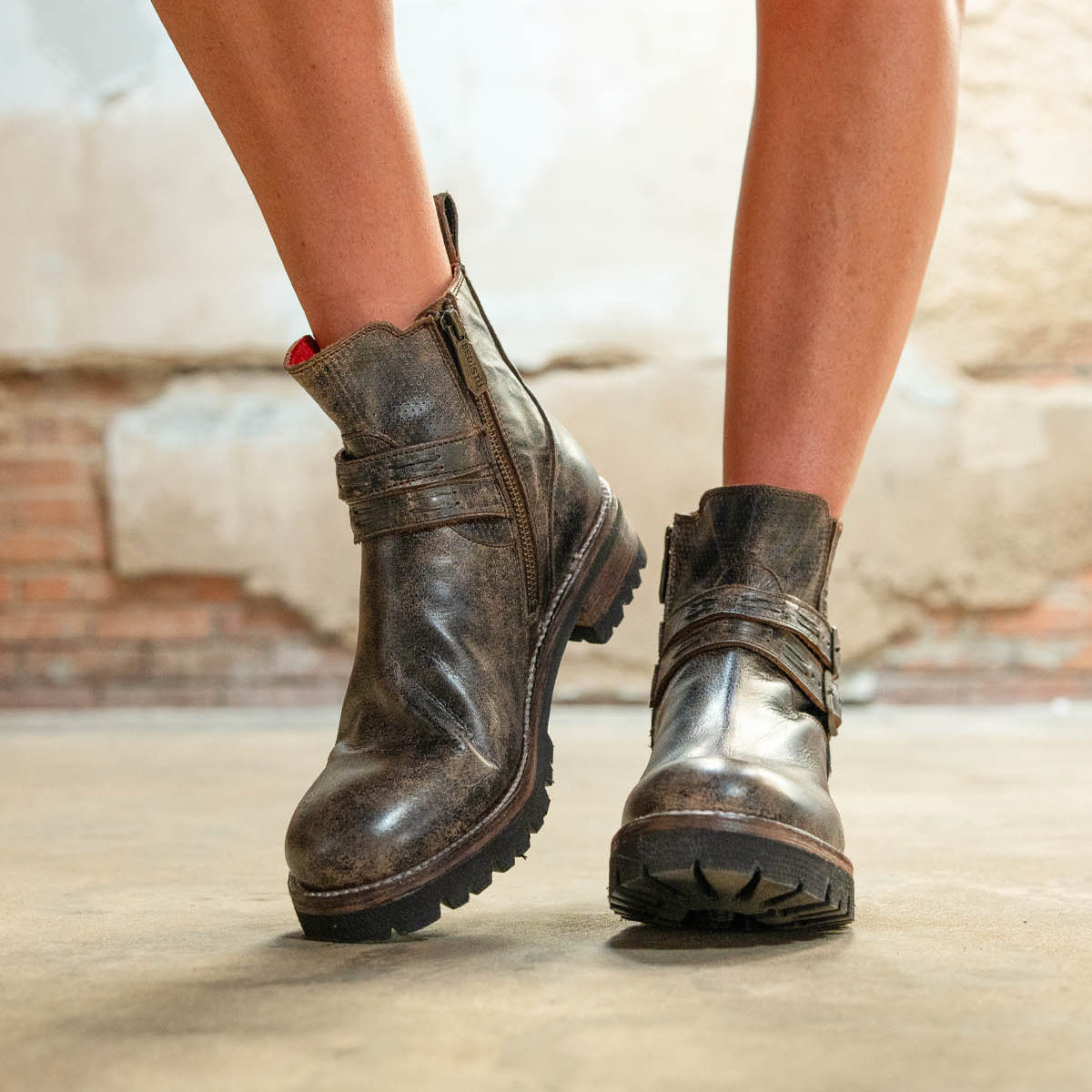 A person wearing the Bed|Stü Daring ankle boots—with worn brown leather, side zippers, and thick soles—stands on a concrete floor against a textured wall, showcasing an effortlessly rugged style that's perfect for everyday wear.