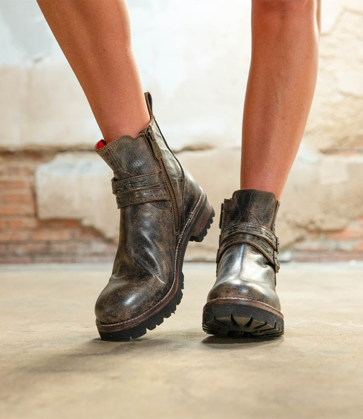 A person wearing the Bed|Stü Daring ankle boots—with worn brown leather, side zippers, and thick soles—stands on a concrete floor against a textured wall, showcasing an effortlessly rugged style that's perfect for everyday wear.