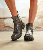 A person wearing the Bed|Stü Daring ankle boots—with worn brown leather, side zippers, and thick soles—stands on a concrete floor against a textured wall, showcasing an effortlessly rugged style that's perfect for everyday wear.