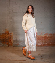 A woman stands indoors against a brick and concrete wall, wearing a cream sweater, a white lace-trimmed skirt, and Bed|Stü's Daring rugged ankle boots.