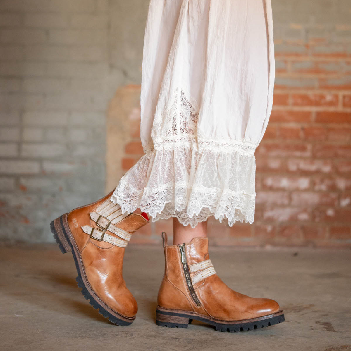 Someone wearing a white lace-trimmed dress paired with Bed|Stü’s Daring brown leather ankle boots, featuring stacked heels, chunky soles, and side zippers, stands indoors against a brick wall.