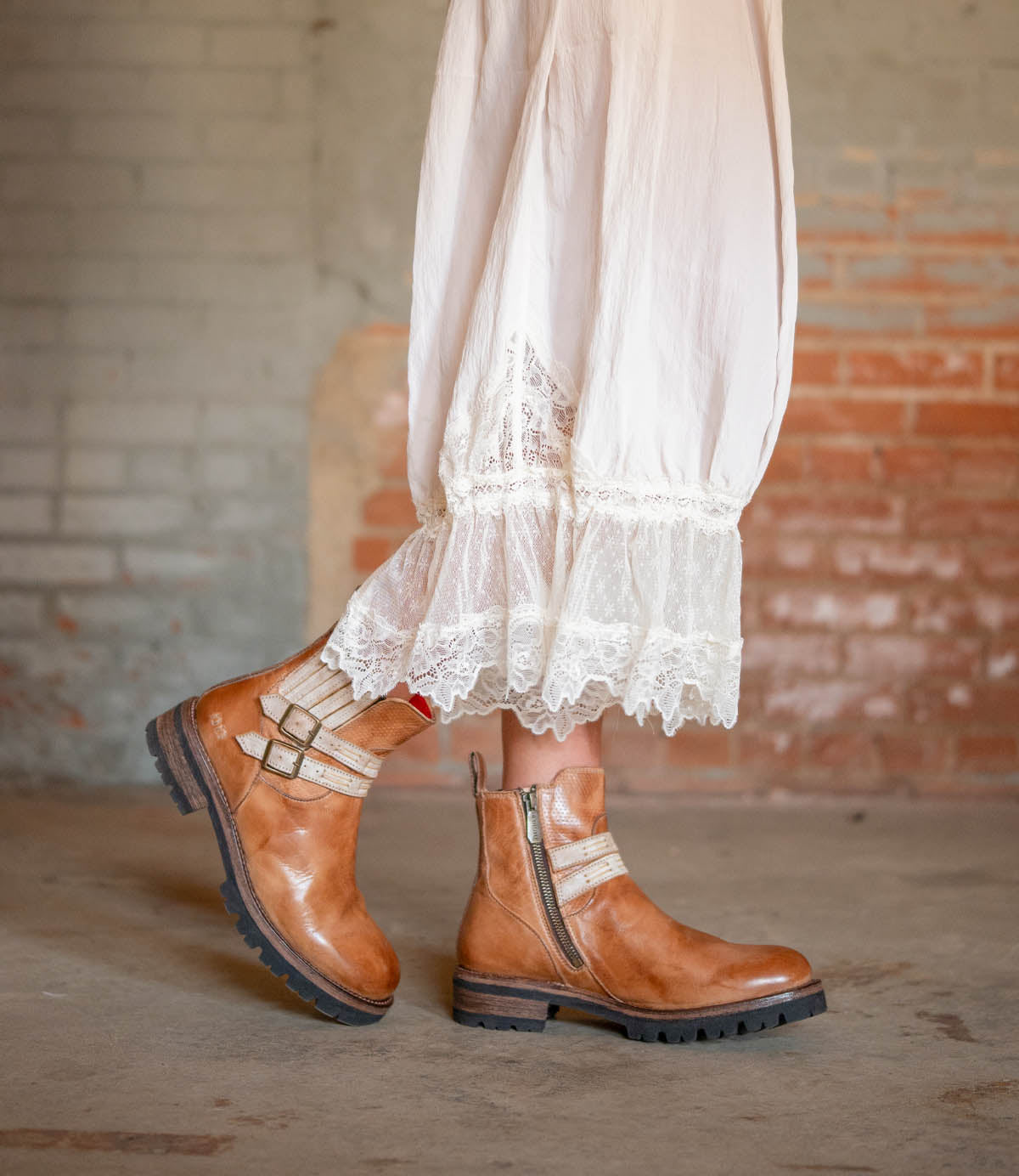 Someone wearing a white lace-trimmed dress paired with Bed|Stü’s Daring brown leather ankle boots, featuring stacked heels, chunky soles, and side zippers, stands indoors against a brick wall.