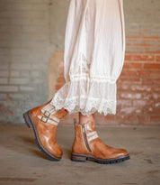 Someone wearing a white lace-trimmed dress paired with Bed|Stü’s Daring brown leather ankle boots, featuring stacked heels, chunky soles, and side zippers, stands indoors against a brick wall.