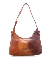 The Denton by Bed|Stü is a brown leather shoulder bag featuring a single strap, visible stitching, and a branded patch on the front. Crafted from vegetable-tanned leather with a zip-top closure, it is displayed against a plain white background.