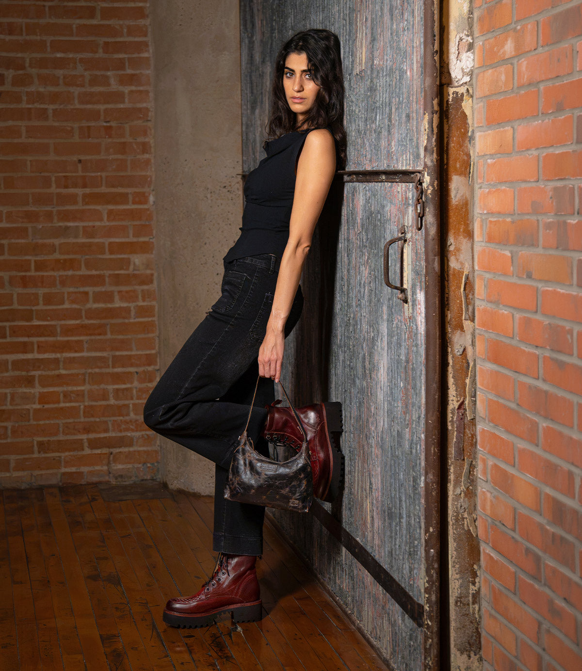 A woman in black clothing and burgundy lace-up boots leans against a large metal door, holding a small, vegetable-tanned Bed|Stü Denton leather handbag.
