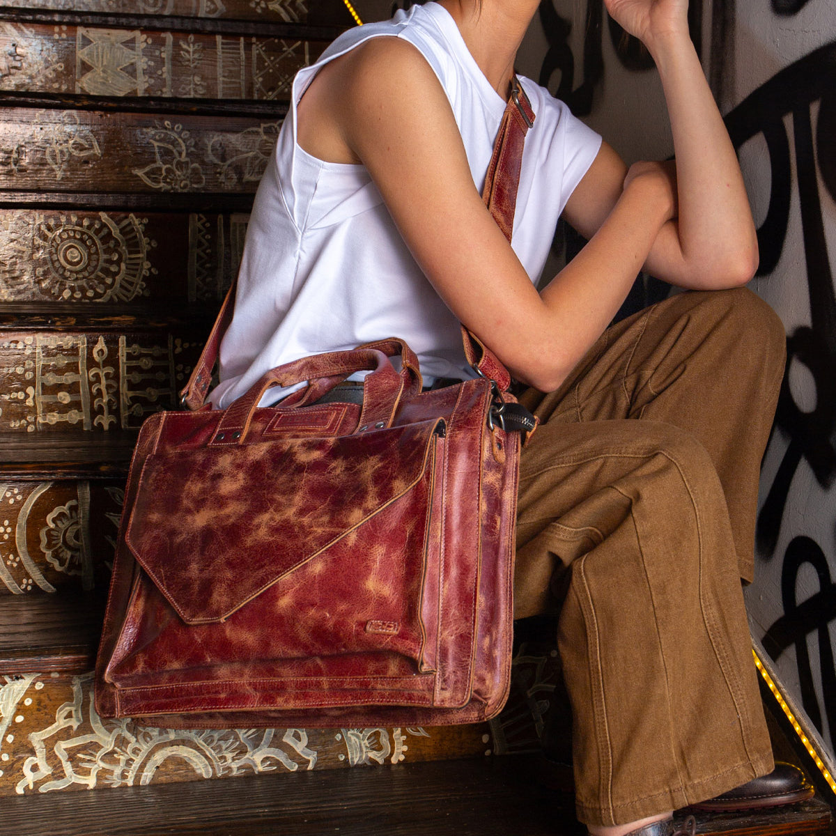 A person sits on decorated stairs carrying the Bed|Stü Depp messenger bag, crafted from brown leather and featuring an adjustable shoulder strap and organized compartments, paired with a sleeveless white top and brown pants.