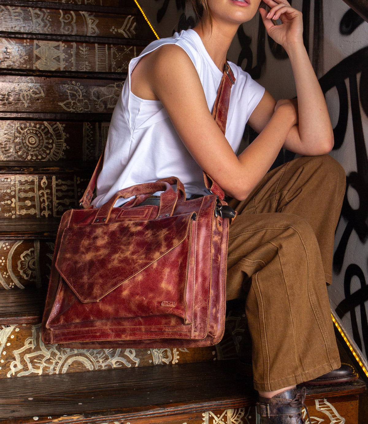 A person sits on decorated stairs carrying the Bed|Stü Depp messenger bag, crafted from brown leather and featuring an adjustable shoulder strap and organized compartments, paired with a sleeveless white top and brown pants.