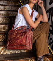 A person sits on decorated stairs carrying the Bed|Stü Depp messenger bag, crafted from brown leather and featuring an adjustable shoulder strap and organized compartments, paired with a sleeveless white top and brown pants.