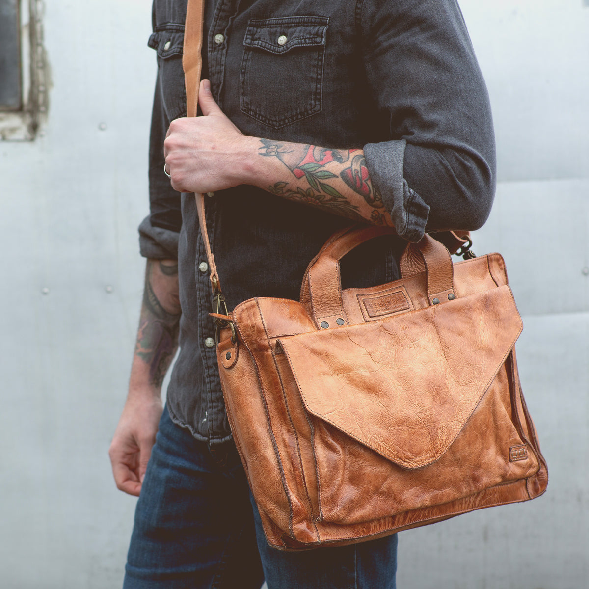 A person wearing a dark shirt and jeans is carrying the Bed|Stü Depp, a tan leather messenger bag with handles and an adjustable shoulder strap, featuring organized compartments for easy storage.