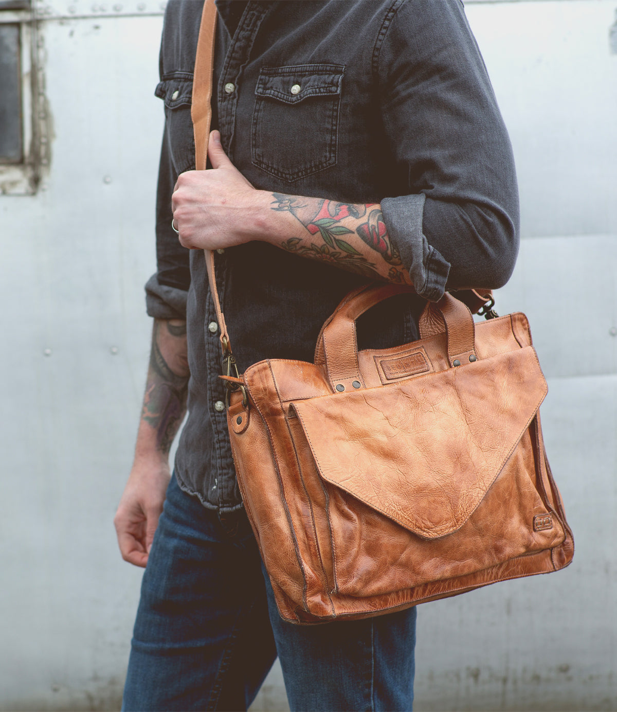A person wearing a dark shirt and jeans is carrying the Bed|Stü Depp, a tan leather messenger bag with handles and an adjustable shoulder strap, featuring organized compartments for easy storage.