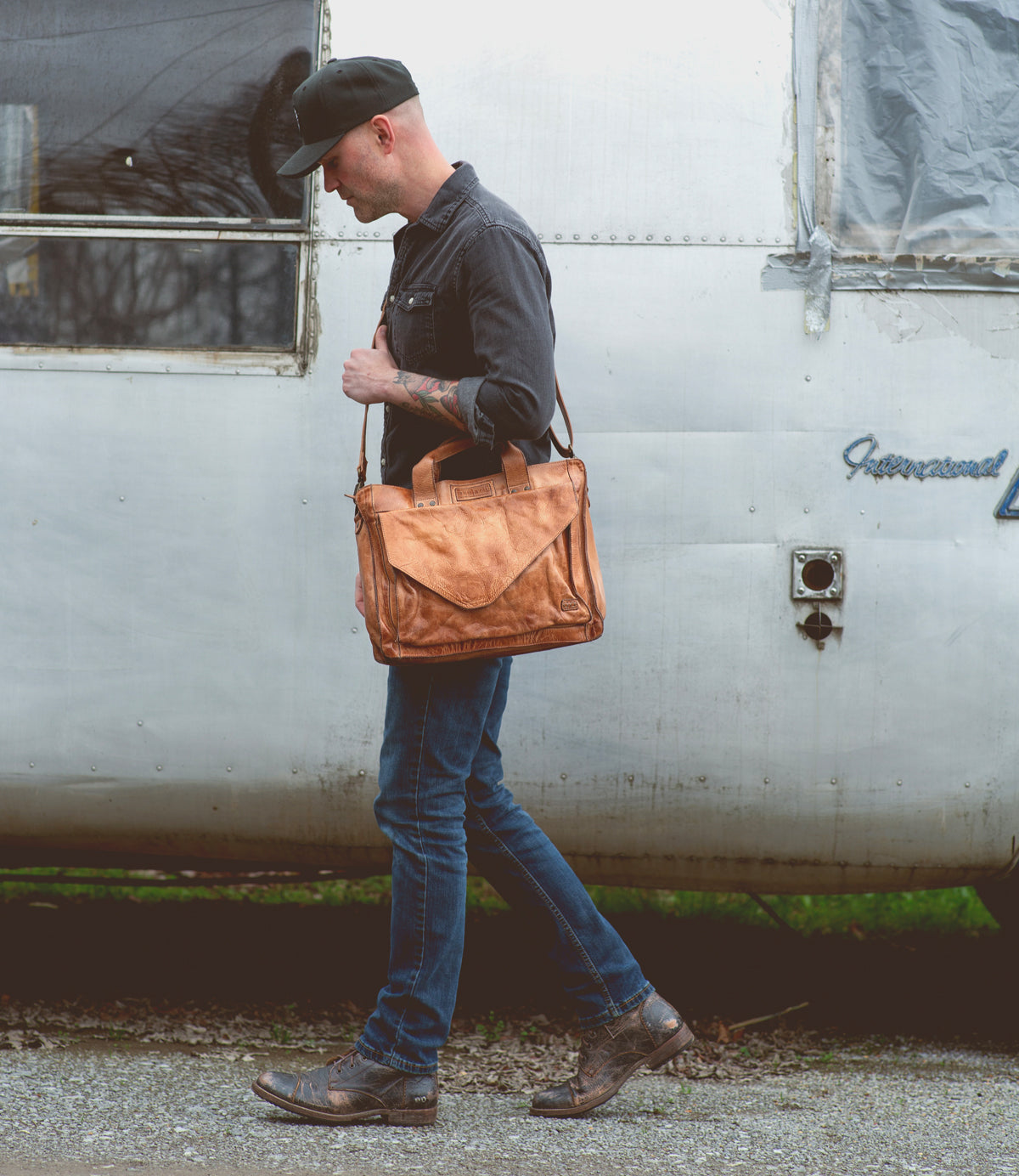 A man in jeans, boots, and a cap walks past a silver trailer while carrying the Bed|Stü Depp brown leather messenger bag with an adjustable shoulder strap over his shoulder.