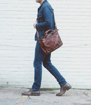 A person in a denim jacket and jeans walks past a white brick wall, carrying the Bed|Stü Depp leather messenger bag with an adjustable shoulder strap and organized compartments, paired with brown shoes.