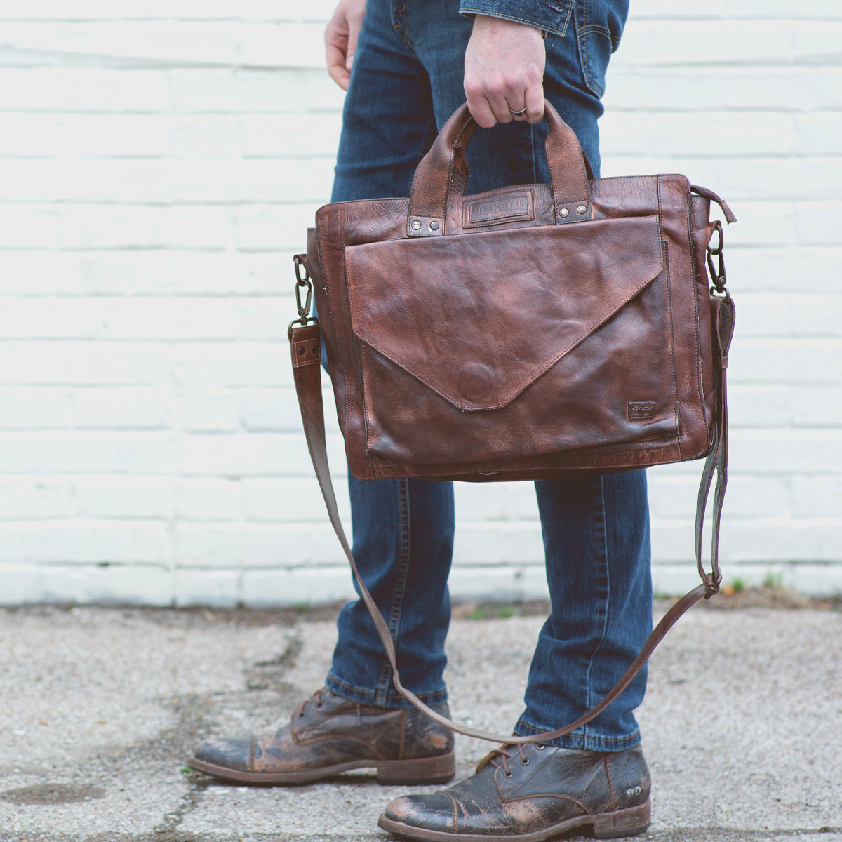 A person stands outdoors, their lower half visible, wearing jeans and carrying the Bed|Stü Depp brown leather messenger bag with organized compartments.