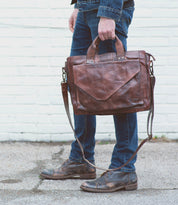 A person stands outdoors, their lower half visible, wearing jeans and carrying the Bed|Stü Depp brown leather messenger bag with organized compartments.