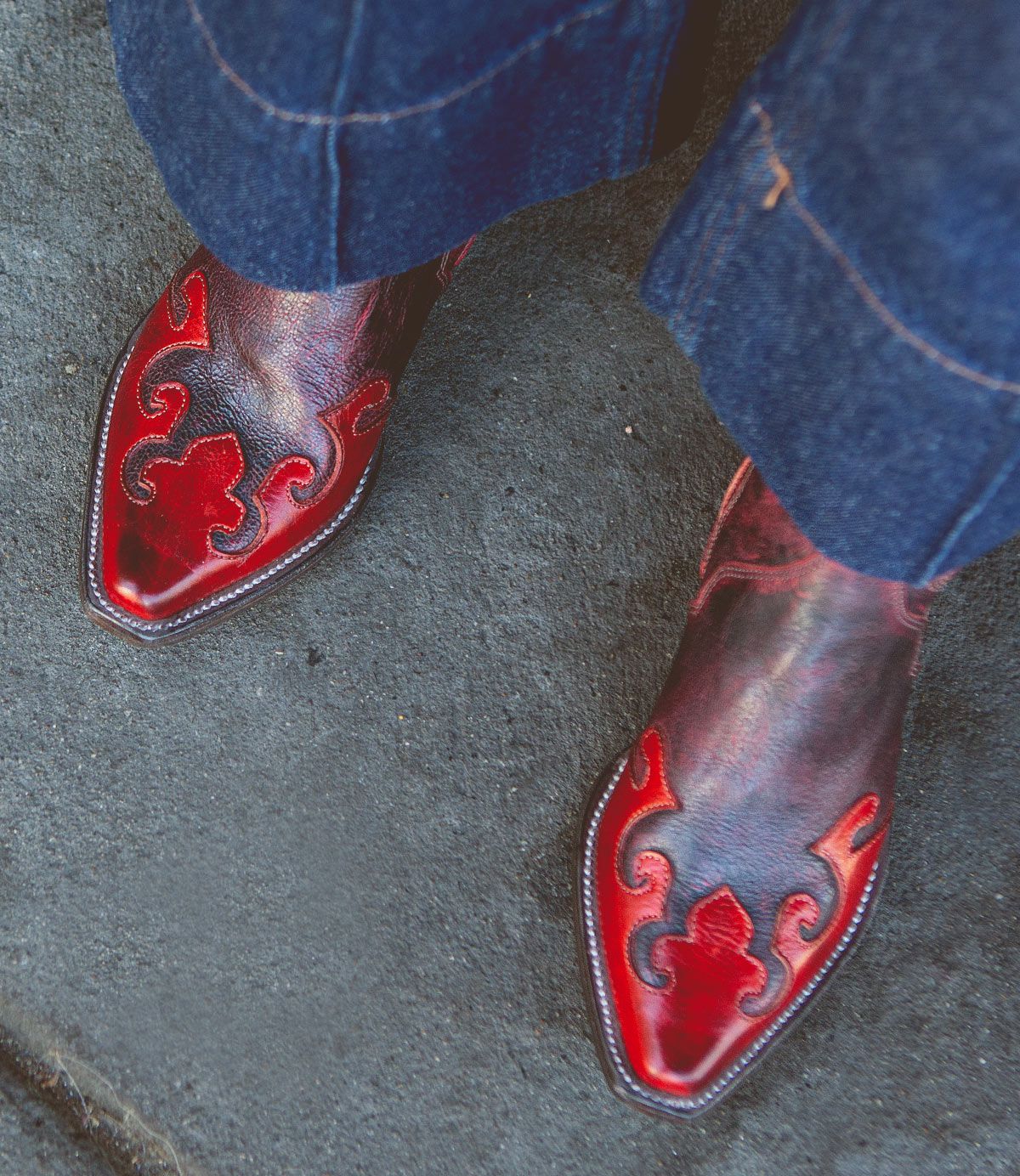 A person wearing blue jeans and Bed|Stü Deuce pointed-toe Western boots, featuring red and black decorative patterns with detailed stitching, stands on a gray concrete surface.