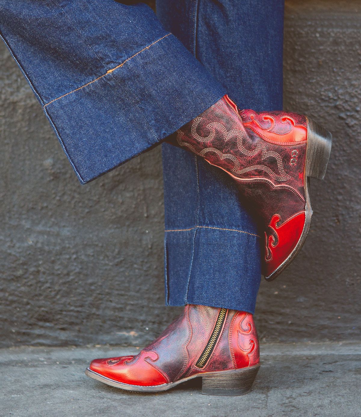 Wearing the Bed|Stü Deuce red Western boots—crafted from vegetable-tanned leather with detailed stitching and side zippers—a person in blue denim pants stands against a dark wall.