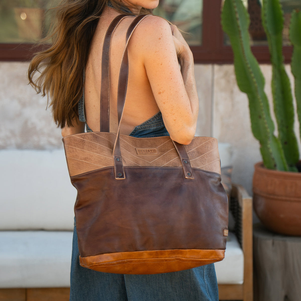 A woman with long hair carries the spacious Bed|Stü Dome brown leather tote over her shoulder, standing outdoors near a cactus and a white outdoor sofa. The roomy interior of the Dome bag is perfect for storing work essentials while on the go.