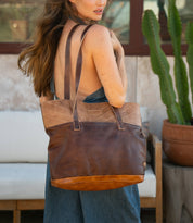 A woman with long hair carries the spacious Bed|Stü Dome brown leather tote over her shoulder, standing outdoors near a cactus and a white outdoor sofa. The roomy interior of the Dome bag is perfect for storing work essentials while on the go.