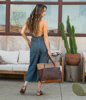A woman stands on a patio near cushioned seating and potted cacti, wearing backless denim overalls and sandals while carrying the Bed|Stü Dome brown leather tote, known for its spacious interior that's perfect for work essentials.