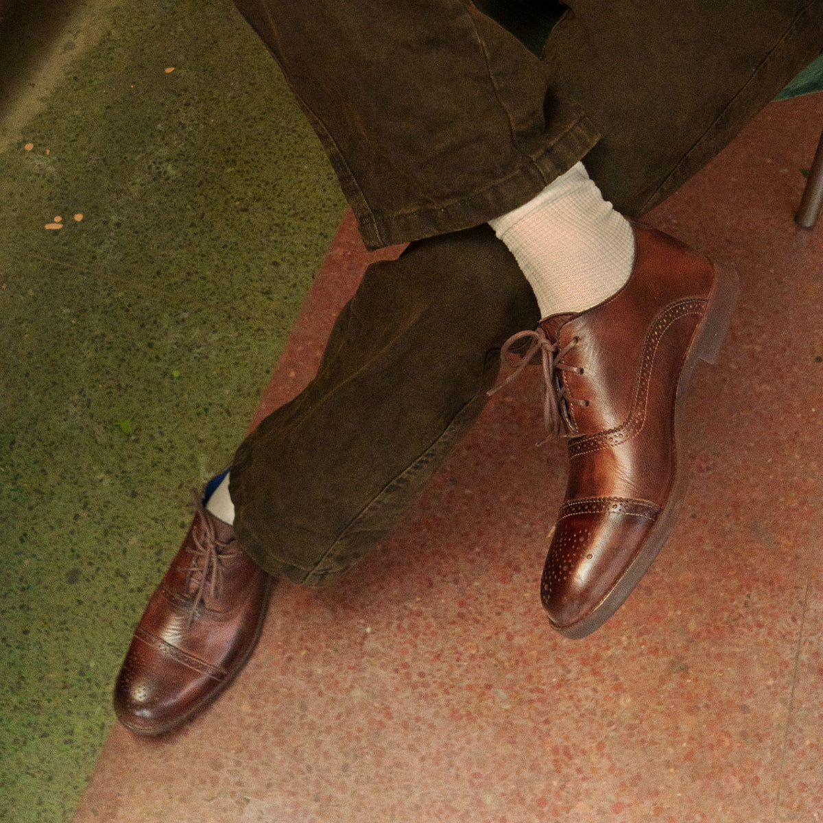 A person wearing Bed|Stü Donatello II brown oxford shoes with a classic cap-toe design, white socks, and brown pants sits with legs crossed on a green and red speckled floor.