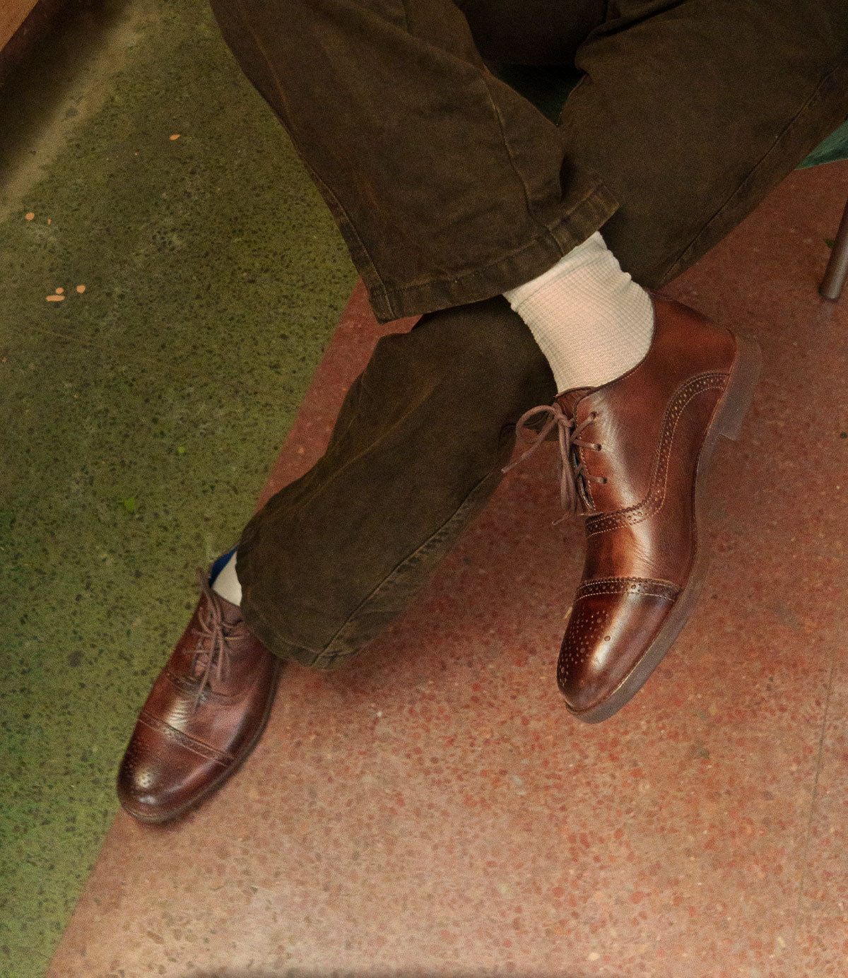 A person wearing Bed|Stü Donatello II brown oxford shoes with a classic cap-toe design, white socks, and brown pants sits with legs crossed on a green and red speckled floor.