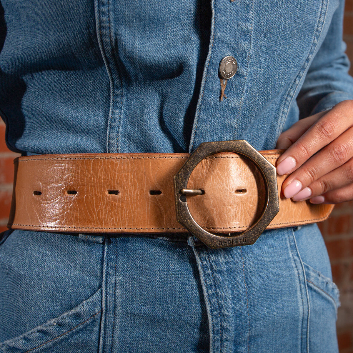 Wearing a blue denim outfit, the person showcases the Bed|Stü Dreampath EM—a wide genuine leather belt with a large round metal buckle—holding the belt with one hand.