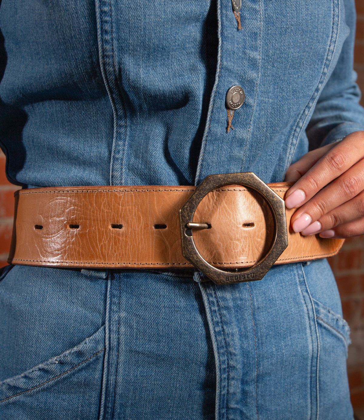 Wearing a blue denim outfit, the person showcases the Bed|Stü Dreampath EM—a wide genuine leather belt with a large round metal buckle—holding the belt with one hand.