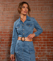 A woman stands against a brick wall, wearing the Bed|Stü Dreampath EM—an elegant blue denim dress featuring long sleeves and a modern-style belt that completes her sophisticated look.