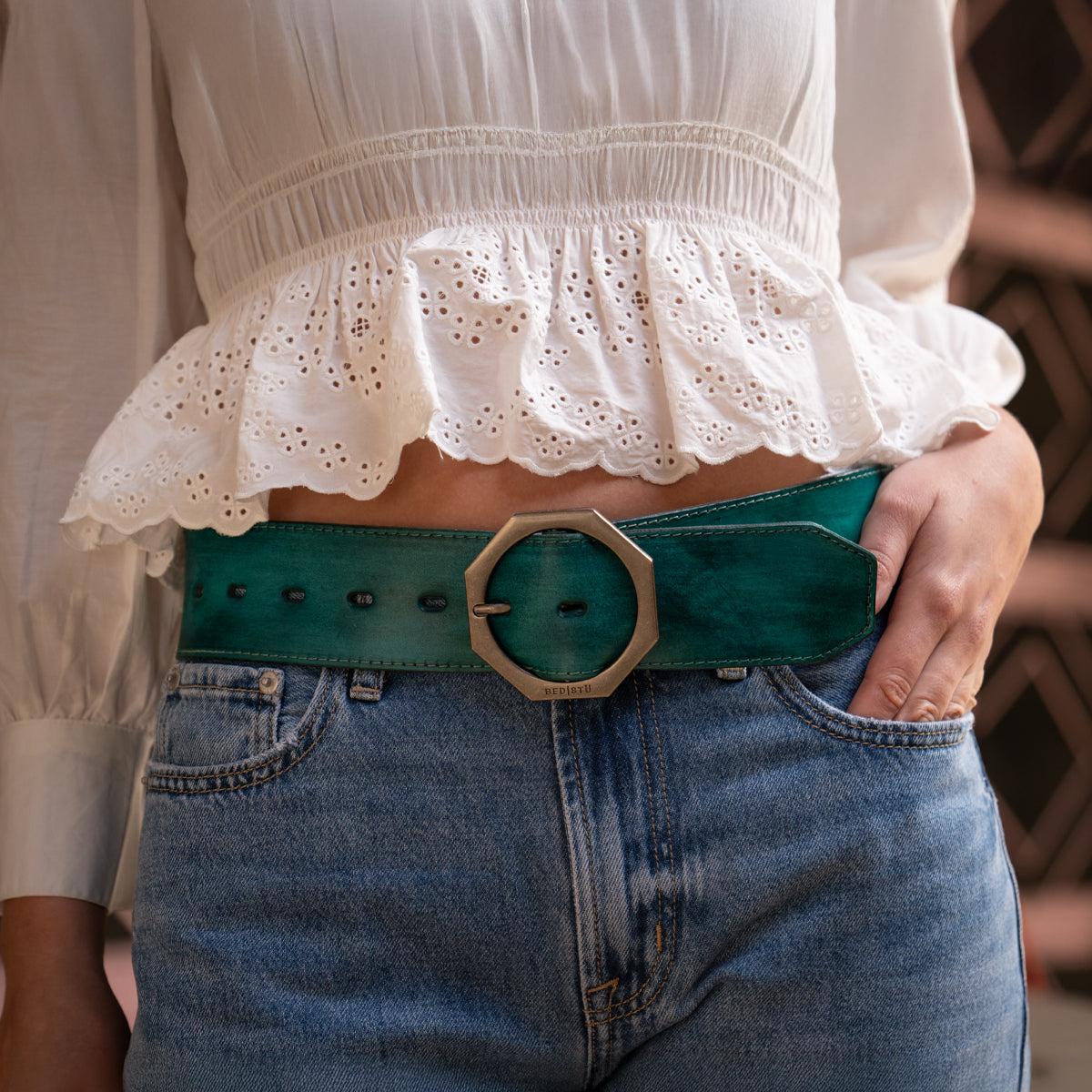 A person wearing a white ruffled blouse, blue jeans, and the Bed|Stü Dreampath wide green genuine leather belt with an octagon metal buckle, standing outdoors.