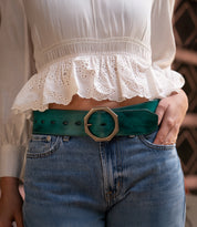 A person wearing a white ruffled blouse, blue jeans, and the Bed|Stü Dreampath wide green genuine leather belt with an octagon metal buckle, standing outdoors.