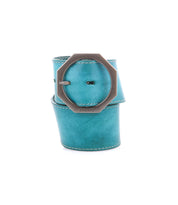 The Bed|Stü Dreampath is a wide turquoise genuine leather belt featuring visible stitching and a large round octagonal metal buckle, artistically rolled up on a white background. This accessory showcases the brand’s commitment to quality craftsmanship and distinctive style.