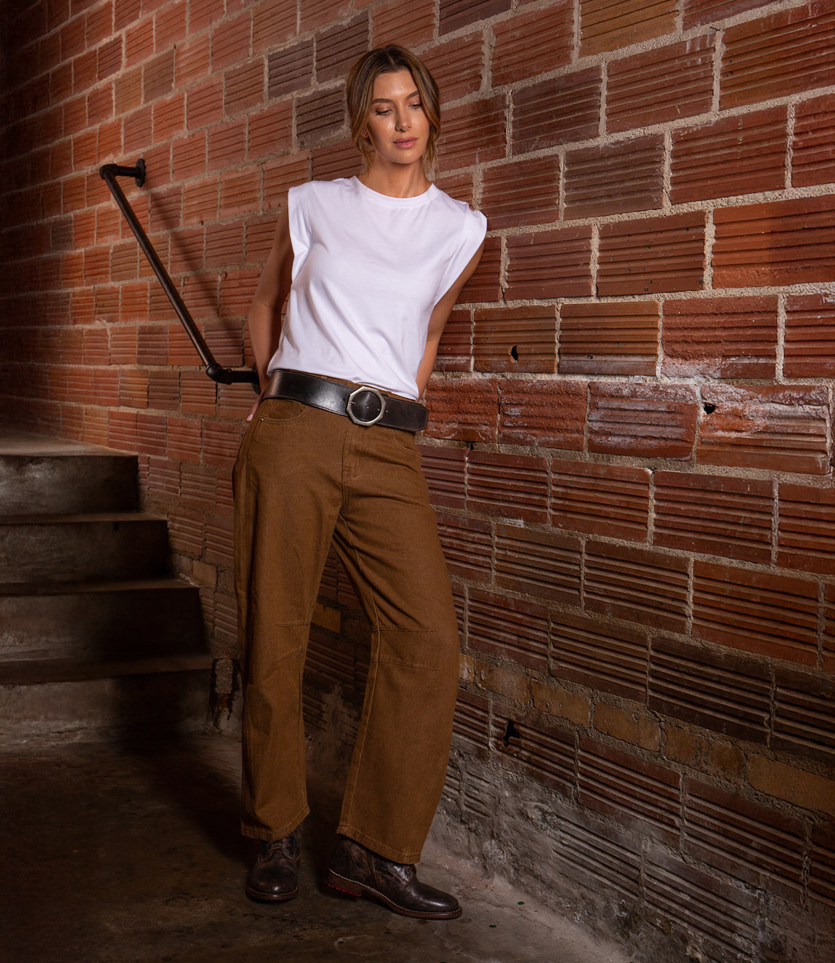 A woman wearing a white sleeveless top and brown pants, accessorized with the Bed|Stü Dreampath genuine leather belt featuring an octagon metal buckle, stands against a brick wall near stairs, looking down with her hands in her pockets.