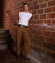 A woman wearing a white sleeveless top and brown pants, accessorized with the Bed|Stü Dreampath genuine leather belt featuring an octagon metal buckle, stands against a brick wall near stairs, looking down with her hands in her pockets.