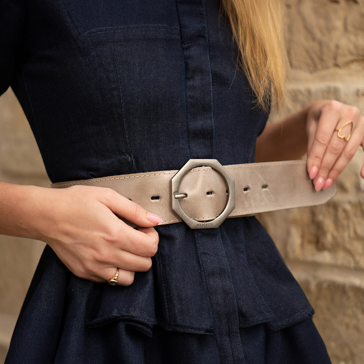 A woman adjusts the Bed|Stü Dreampath genuine leather belt with an octagon-shaped buckle over a dark blue dress, standing against a stone wall.
