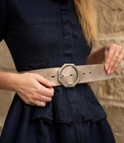 A woman adjusts the Bed|Stü Dreampath genuine leather belt with an octagon-shaped buckle over a dark blue dress, standing against a stone wall.