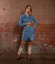 A woman poses against a brick wall wearing a blue denim dress, the Bed|Stü Dreampath genuine leather belt featuring an octagon metal buckle, and two-tone cowboy boots, with one hand on her hip and the other resting by her side.