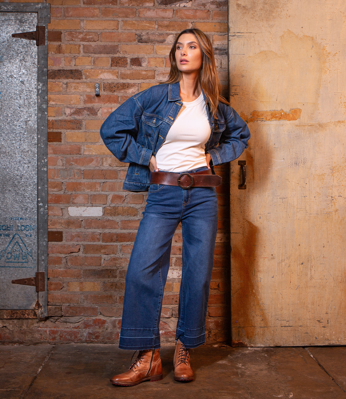 A woman stands against a brick wall wearing a denim jacket, white shirt, wide-leg jeans, and brown Bed|Stü Dreampath boots. She completes her look with a genuine leather belt featuring an octagon metal buckle, hands on hips and gazing slightly to the side.