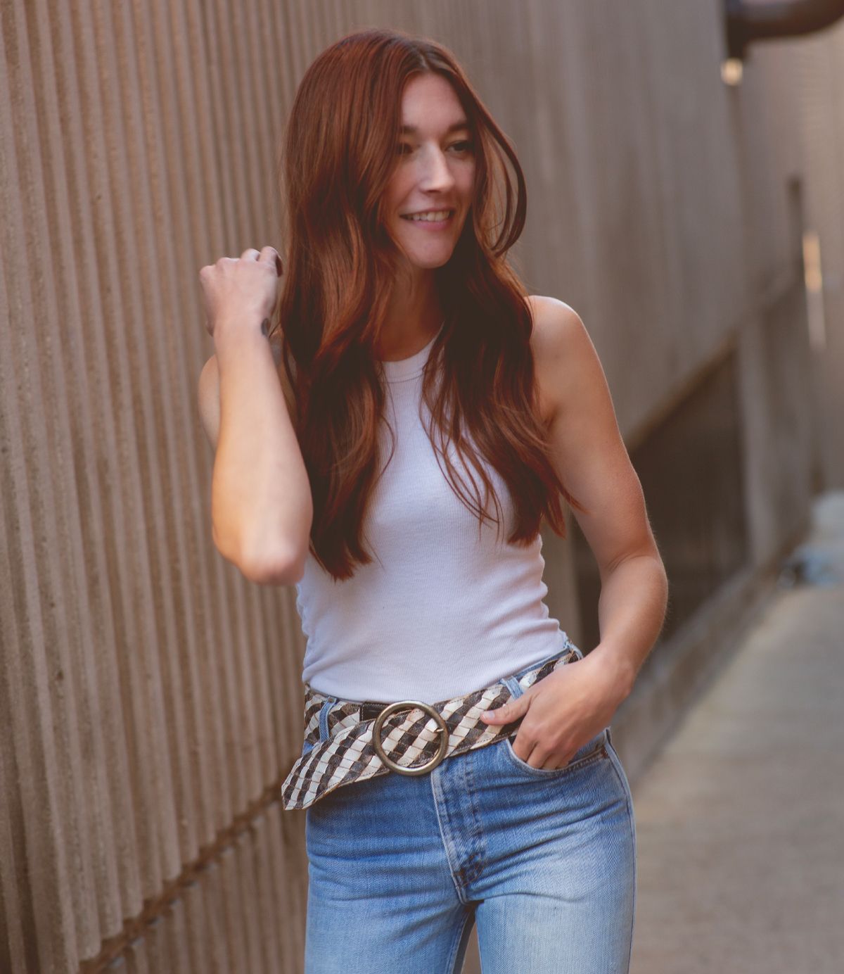 Wearing the Dreamweaver woven leather belt by Bed|Stü, a person with long auburn hair in a white sleeveless top and jeans stands outdoors, smiling with one hand in their pocket—the artisan craftsmanship of the belt perfectly accentuating their bohemian style.